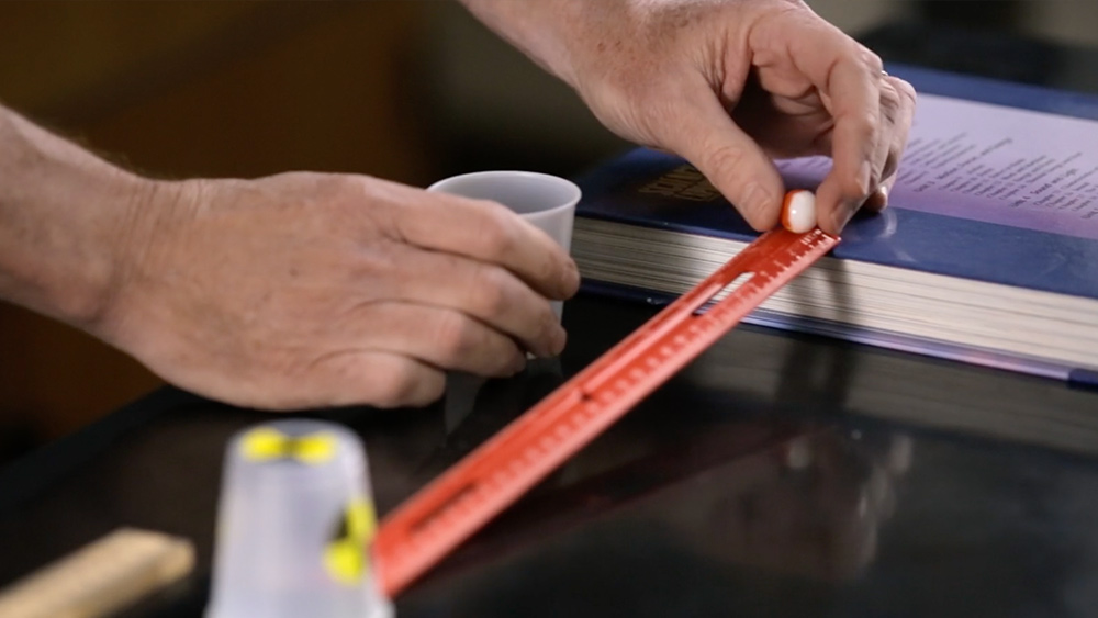 A hand holds a marble on a downward-slanting ruler. 